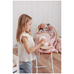 Girl feeding a doll in a pink Asi Dolls high chair with a ruffled bib, other dolls in the background.