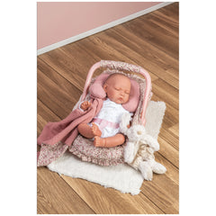 Sleeping doll in a floral Maxi-Cosi with a pink neck cushion and blanket on a wooden floor.