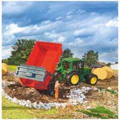 Red Bruder tipping trailer with automatic tailgate attached to a green tractor in a farm setting with hay bales.