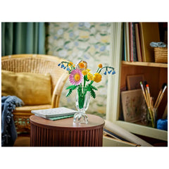 Colorful LEGO® Botanicals Petite Sunny Bouquet set displayed in a clear vase on a round wooden table, surrounded by a cozy living room.