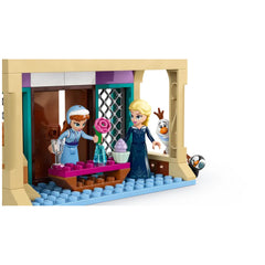 Elsa and Anna mini-dolls stand in a colorful castle room. Anna holds a flower, while Elsa displays a dessert on a table. Olaf peeks through the door.