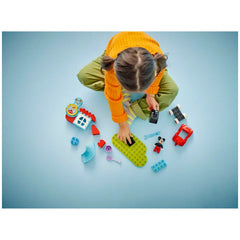 A child in an orange sweater and green pants sits on a blue surface, playing with LEGO® DUPLO® Mickey Mouse Clubhouse pieces, including a green baseplate, a red car, and colorful accessories.