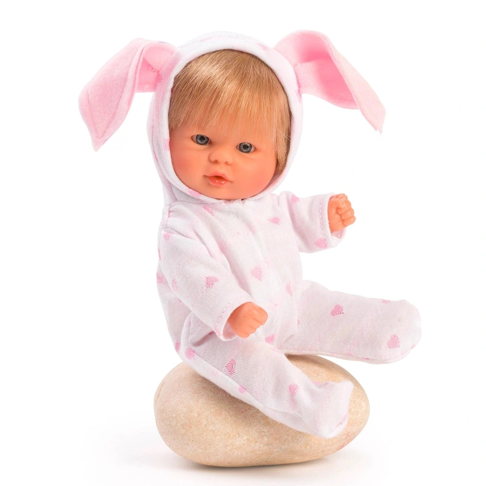 Asi Dolls Bomboncín, 20 cm baby doll with blonde hair in pink-eared pajamas, seated on a stone.
