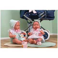 Two ASI Dolls in gingham and lace dresses sit on a rug with wooden blocks, beside a navy blue stroller.