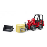 Red Bruder SchΓ€ffer Compact Loader 2630 toy with bale gripper holding yellow bale, detachable bucket nearby.