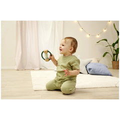 Eichhorn Baby HiPP Sound Rattle is held by a smiling baby in a green outfit, sitting on a white rug in a cozy indoor setting.