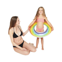 Rainbow swimming ring held by a smiling girl in a pink bikini, with a woman in a black bikini sitting nearby on a white surfa