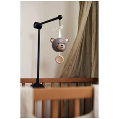 Jollein musical hanger features a knitted teddy bear hanging from a beige fabric draped over a brown wooden crib railing.