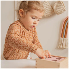 Child with blonde hair in an orange leopard print shirt holds a wooden shape-sorting box on a smooth white table.