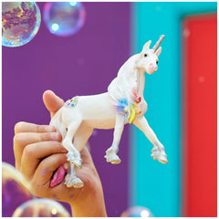 Rainbow Love Unicorn Stallion figurine held in a hand against a vibrant gradient background with floating bubbles.