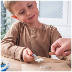 A young child in a brown shirt is focused on a brown object at a light wooden table with a blue bowl.