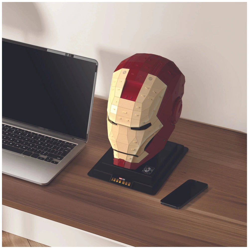 Iron Man-themed figurine with red helmet and yellow details stands on a black stand atop a wooden desk with a laptop and smar
