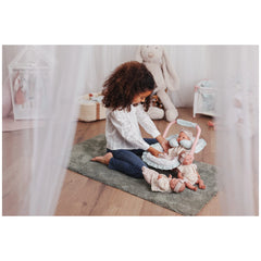 Child playing with ASI Dolls Oli Romper, seated on a gray rug in a cozy room, surrounded by plush toys and doll accessories.