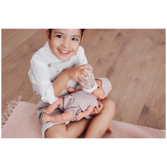 Child in white overalls feeding a doll with a floral bottle holder, sitting on a pink mat.