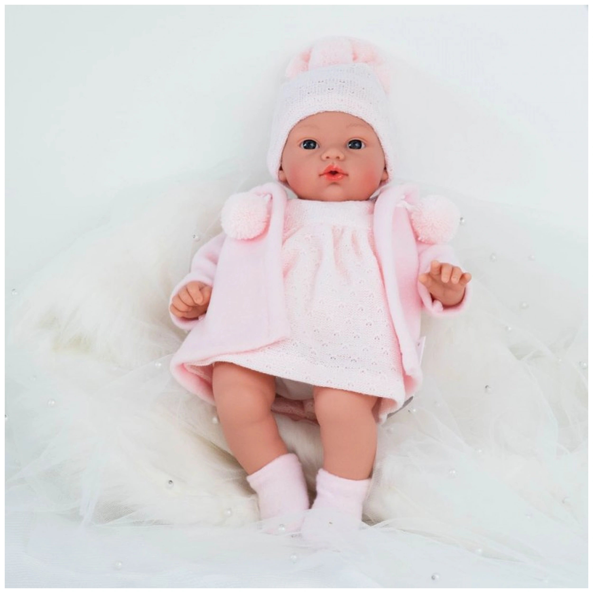 Koke doll in pink duffle coat and hat, lying on soft white fabric, showcasing realistic features and blue eyes.