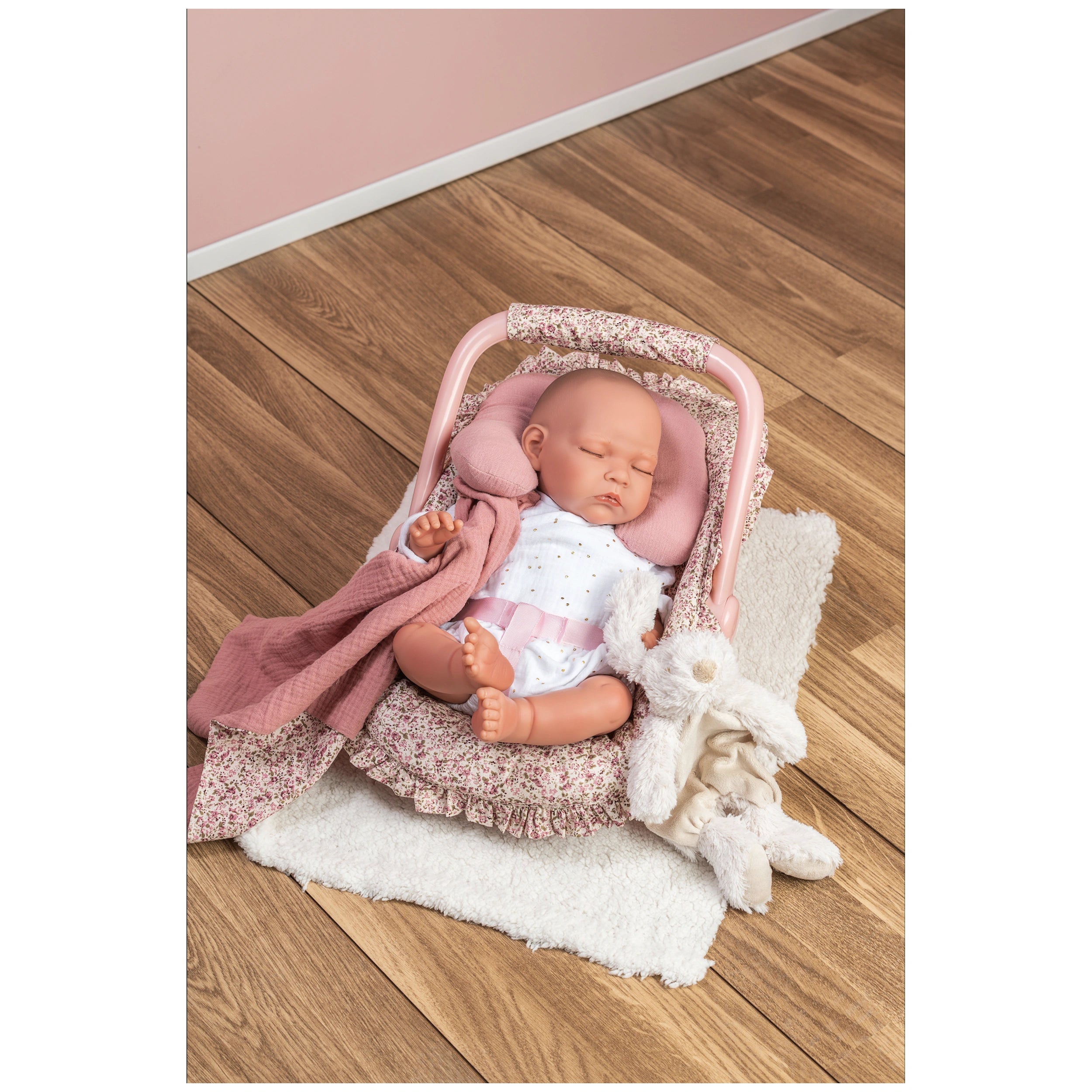Sleeping doll in a floral Maxi-Cosi with a pink neck cushion and blanket on a wooden floor.
