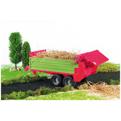 Red and green toy dung spreader with movable parts, loaded with straw, on a miniature farm scene with grass and trees.