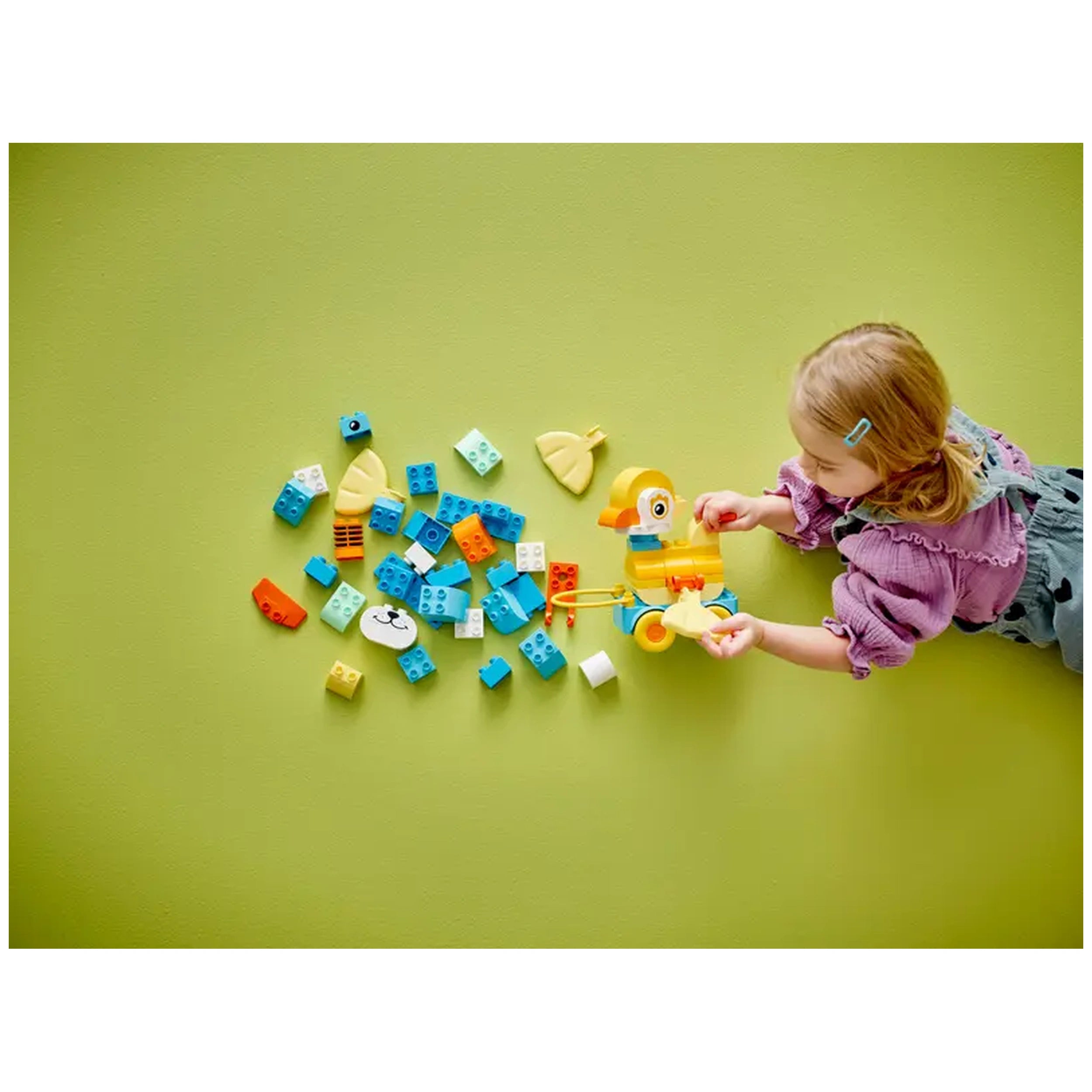 A child with blonde hair plays on a green surface, constructing a yellow duck with colorful LEGO pieces scattered around.