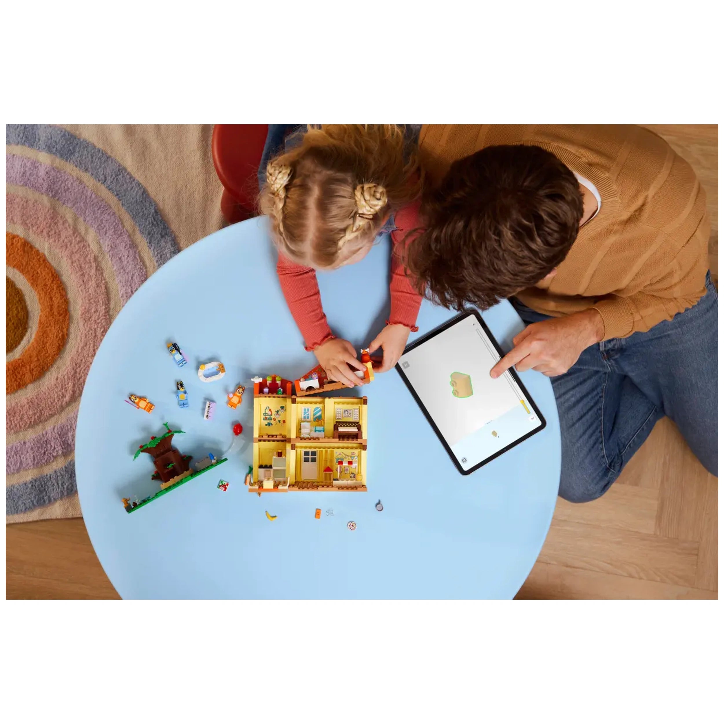 A young child and an adult are engaged in play at a light blue table, focusing on the LEGO Bluey’s Family House set. The child is assembling characters and accessories, while the adult uses a tablet for guidance. Surrounding them are scattered playset pieces and a colorful rug underneath.