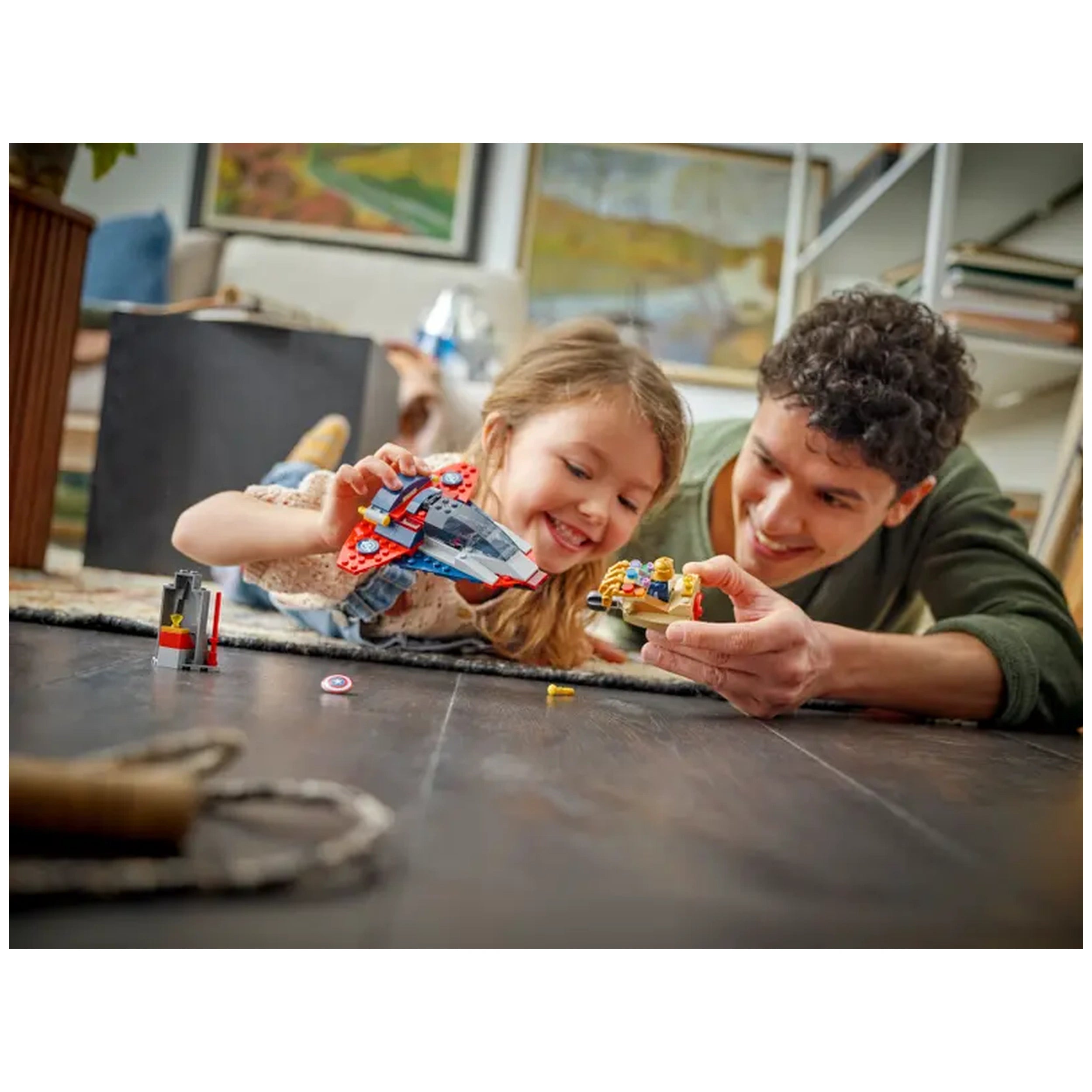 A young girl and an adult play on a wooden floor, smiling as they engage with LEGO action figures, including Captain America and a spaceship, surrounded by colorful building pieces.