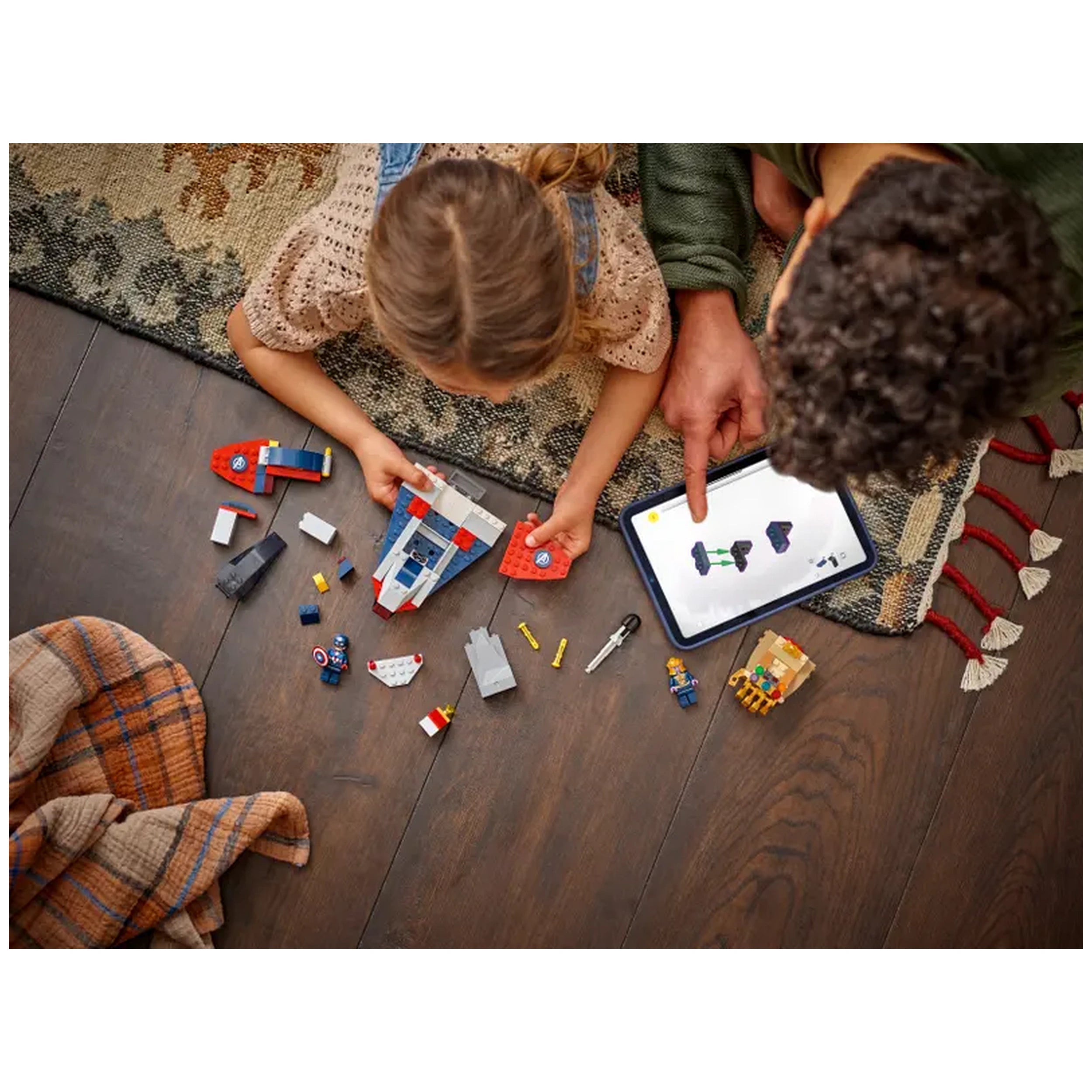 A child builds with LEGO pieces, holding a red and blue spaceship while an adult points to a tablet displaying construction instructions. Various colorful blocks and minifigures are scattered on a wooden floor beside them. A cozy blanket is draped nearby.