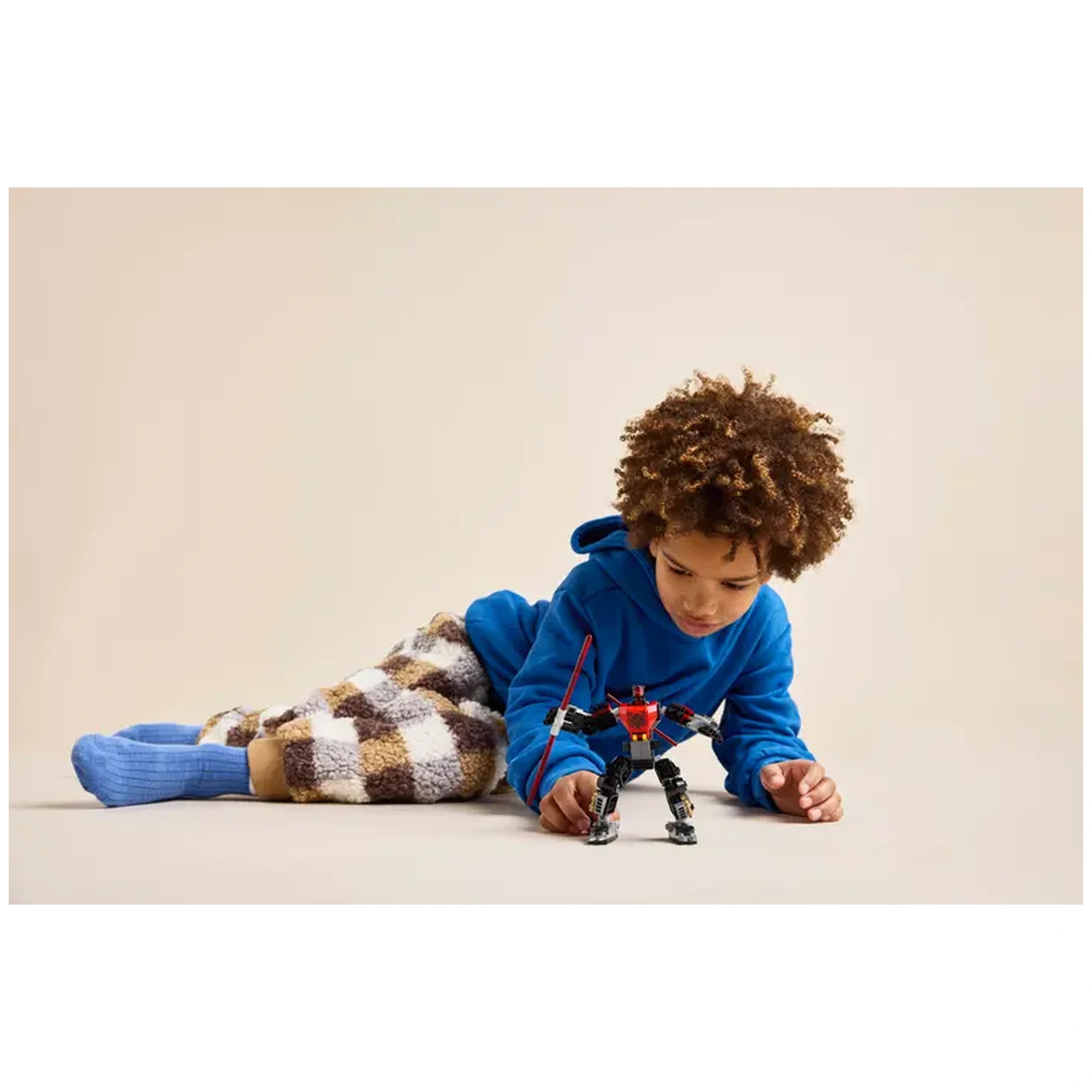 Young child in a blue hoodie, lying on a beige surface, focused on playing with a LEGO Darth Maul Mech featuring posable limbs and a red Lightsaber.