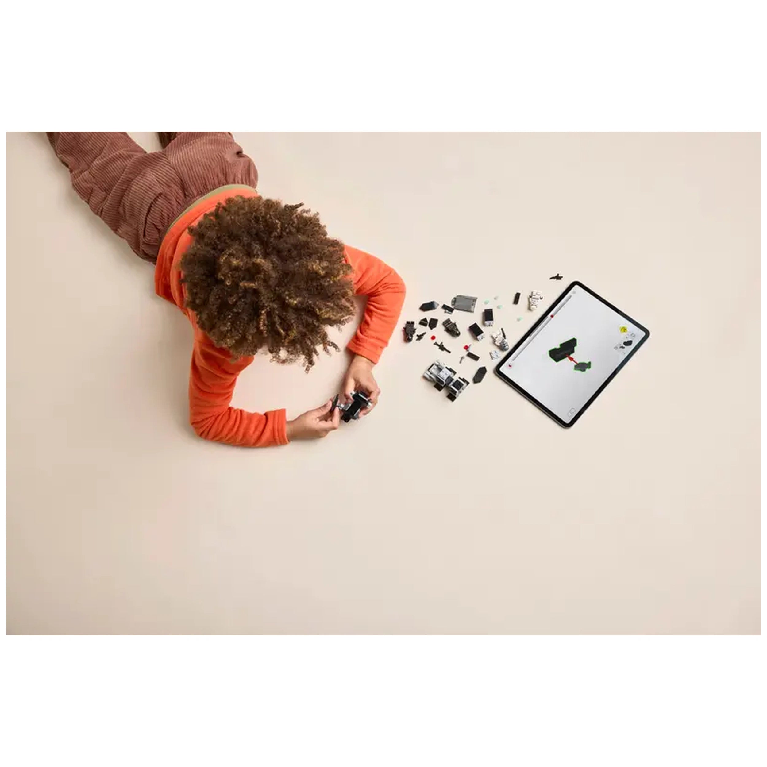 Child in an orange shirt assembling LEGO pieces on a light-colored surface, surrounded by scattered bricks and a tablet showing instructions.