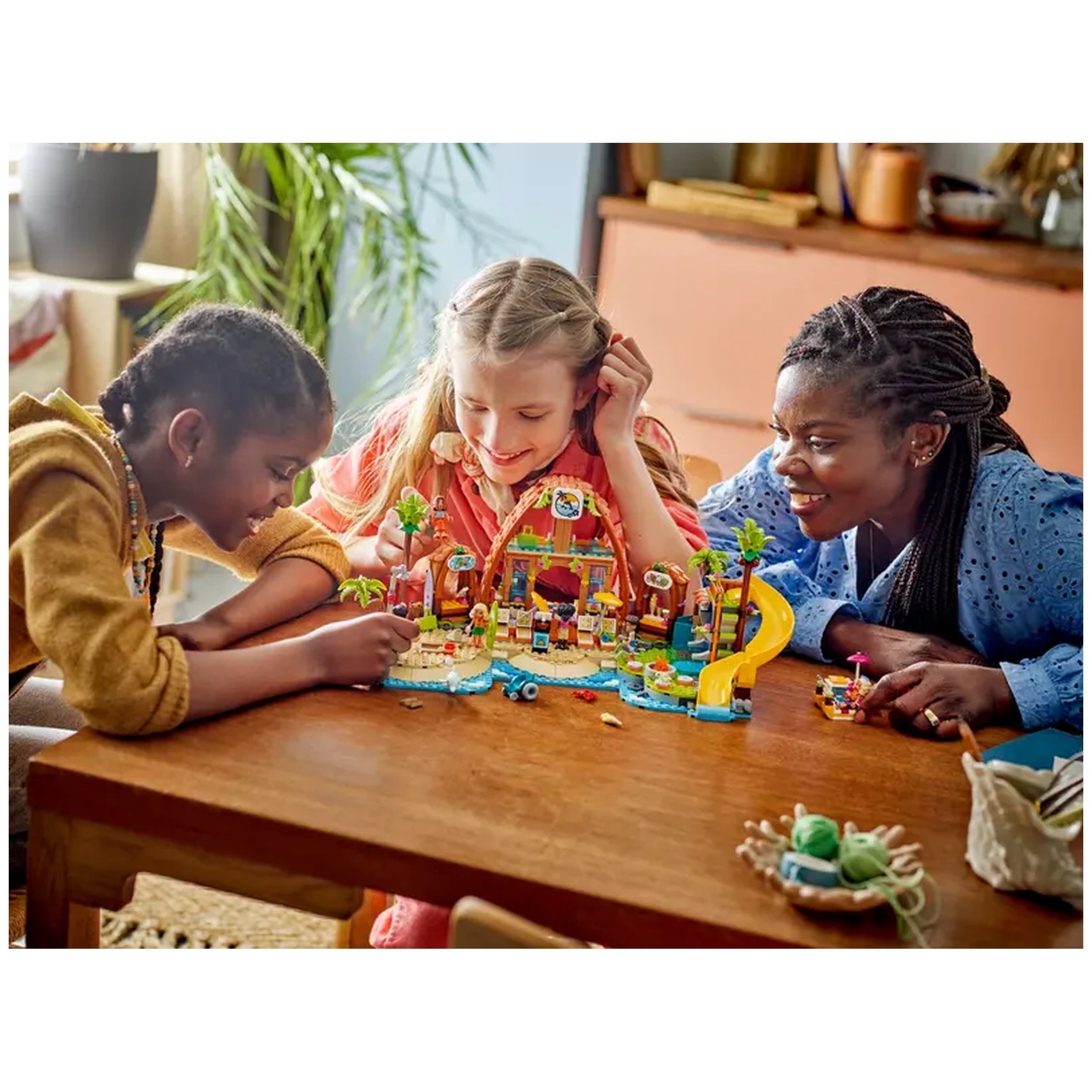 Three children play together at a wooden table, immersed in building the LEGO Family Vacation Beach Resort set. The scene includes a vibrant beach with palm trees, a slide, and minidolls, showcasing creativity and joy. Sunlight brightens the room, enhancing the atmosphere of fun and imagination.