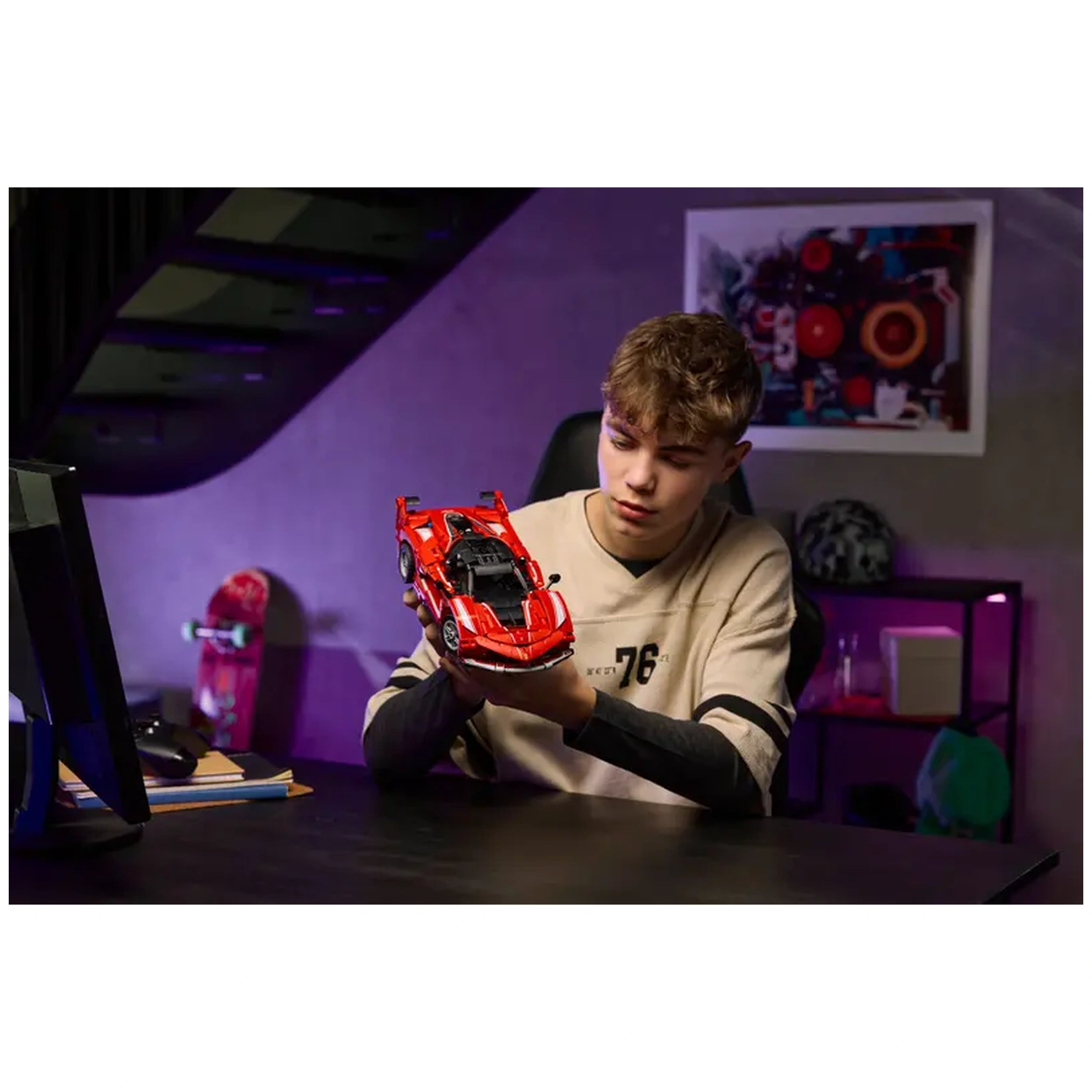A young boy carefully examines a bright red LEGO Ferrari FXX K model car, seated at a dark desk with a computer and books nearby. The background features a purple wall and a colorful artwork, creating a vibrant atmosphere.