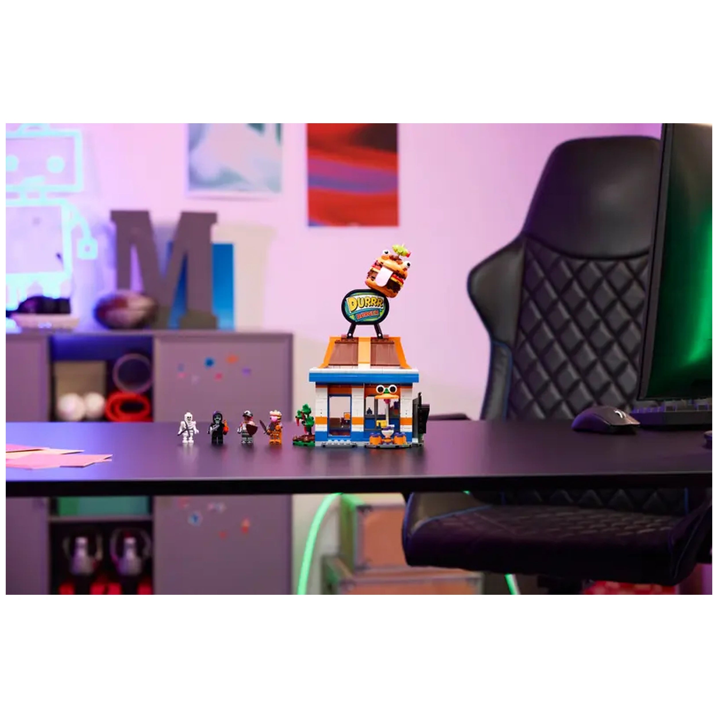 LEGO Fortnite Durrr Burger Restaurant set on a black table, featuring colorful burger-shaped roof, condiment station, and five toy figures lined up in front. A gaming chair and vibrant background add to the playful scene.