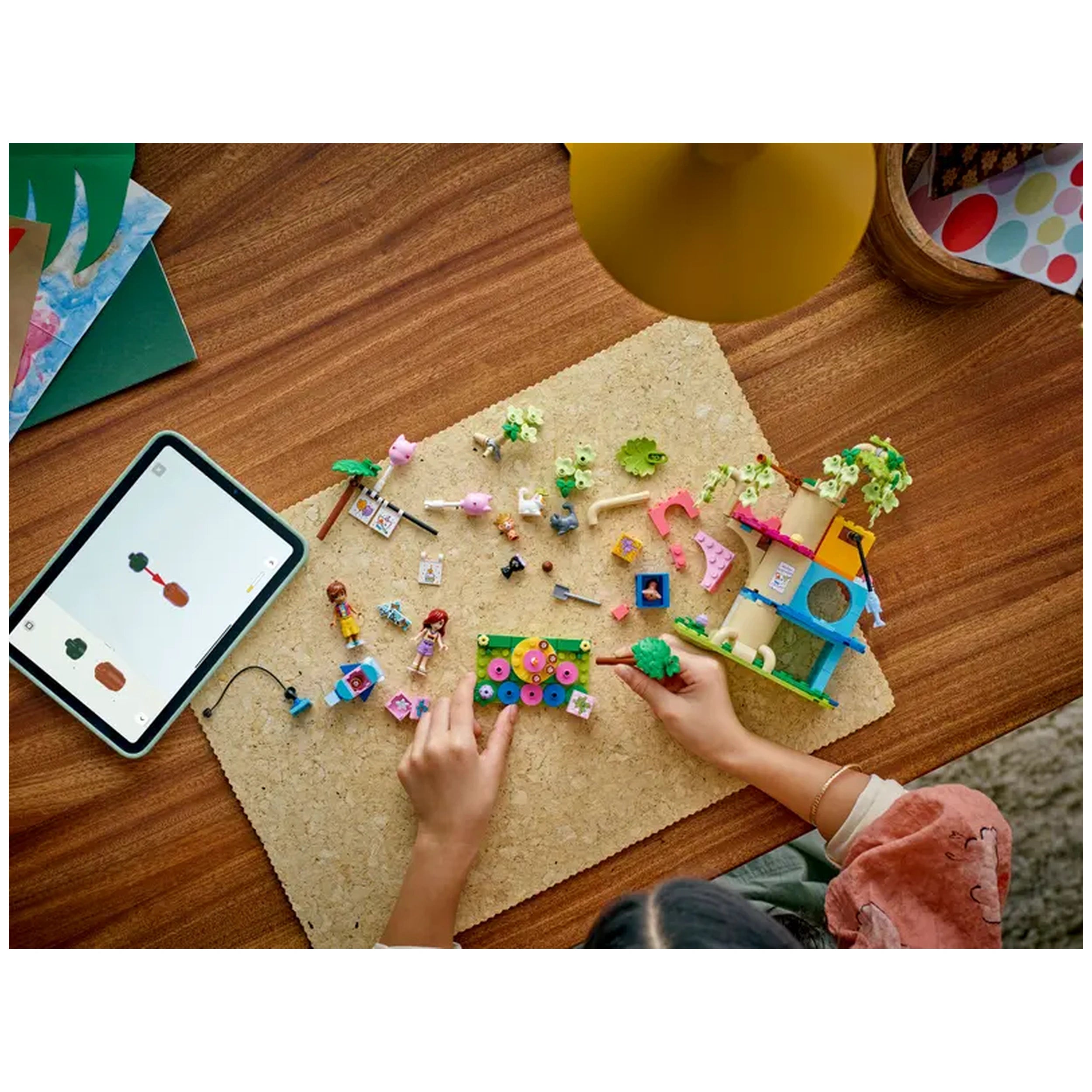 Colorful LEGO Friends set on a cork mat, featuring two minidolls, cute cat figures, birthday decorations, and a play tree house, with hands assembling pieces and a tablet displaying instructions.