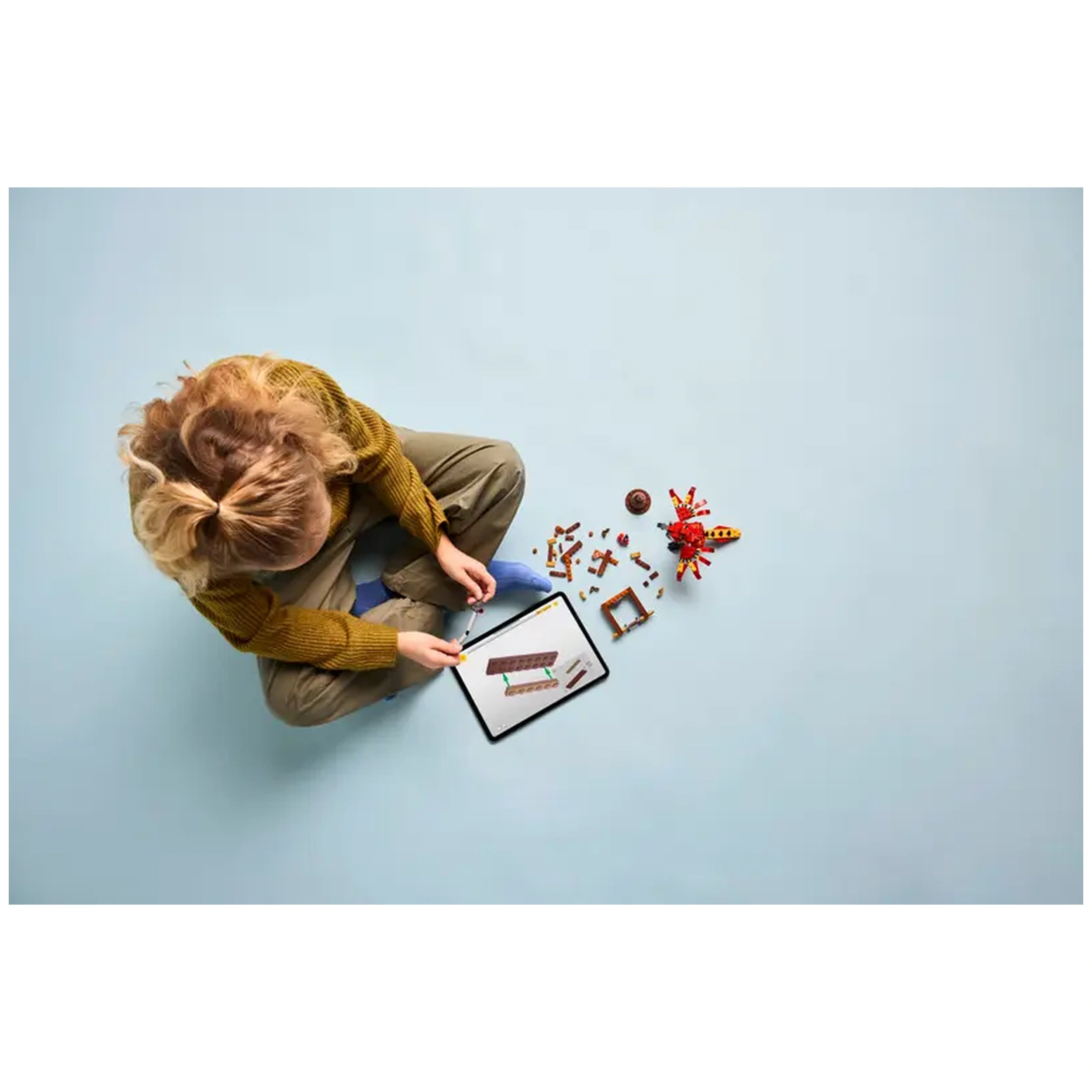 A child sits on a light blue floor, focused on building a LEGO model with various colorful pieces nearby. An iPad displays assembly instructions while the child holds a LEGO piece, wearing a cozy yellow sweater and grey pants.
