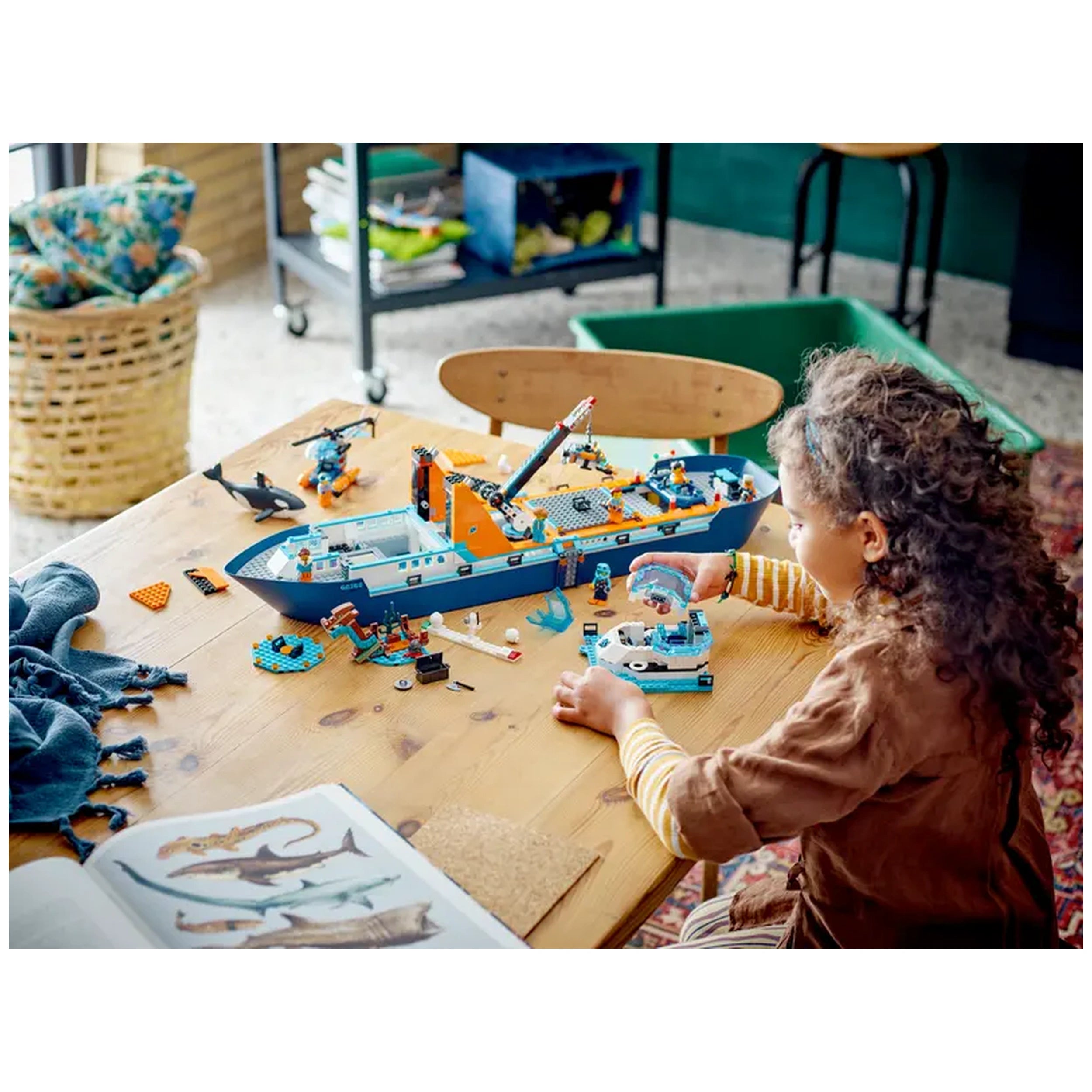 A child with curly hair builds the LEGO City Arctic Explorer Ship on a wooden table, surrounded by colorful toy pieces and ocean-themed miniatures.
