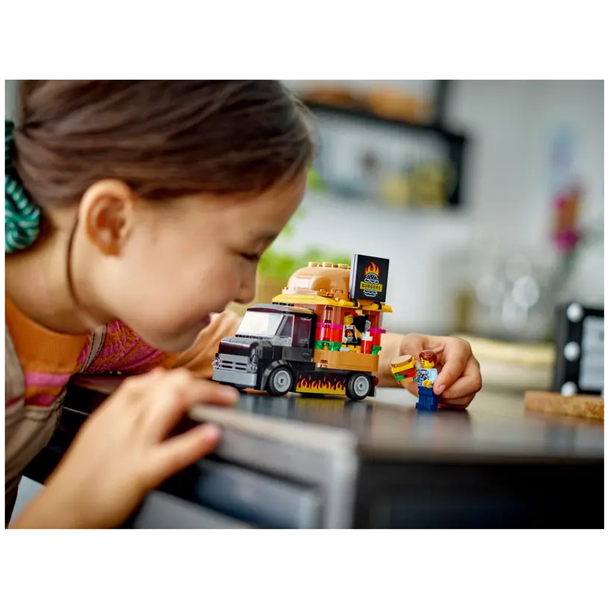 A young girl leans over a colorful LEGO® City Burger Truck, playfully serving a burger to a minifigure. The truck features a giant burger on top and vibrant food details.