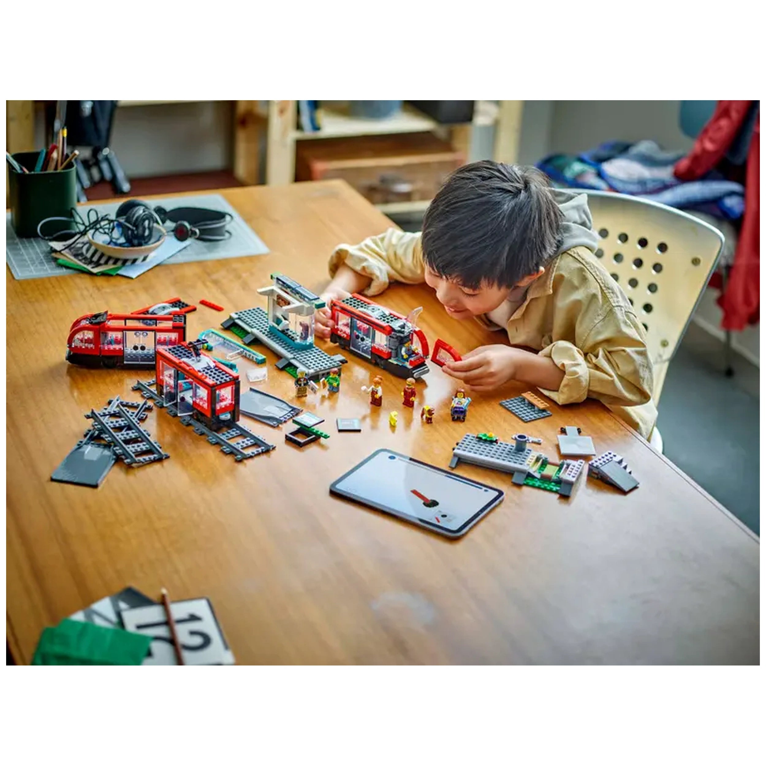 A young boy is assembling the LEGO® City Downtown Streetcar and Station set on a wooden table. Colorful streetcars, minifigures, and tracks are scattered around him, alongside a tablet and stationary items. He focuses intently on connecting pieces, showcasing creativity and engagement in imaginative play.