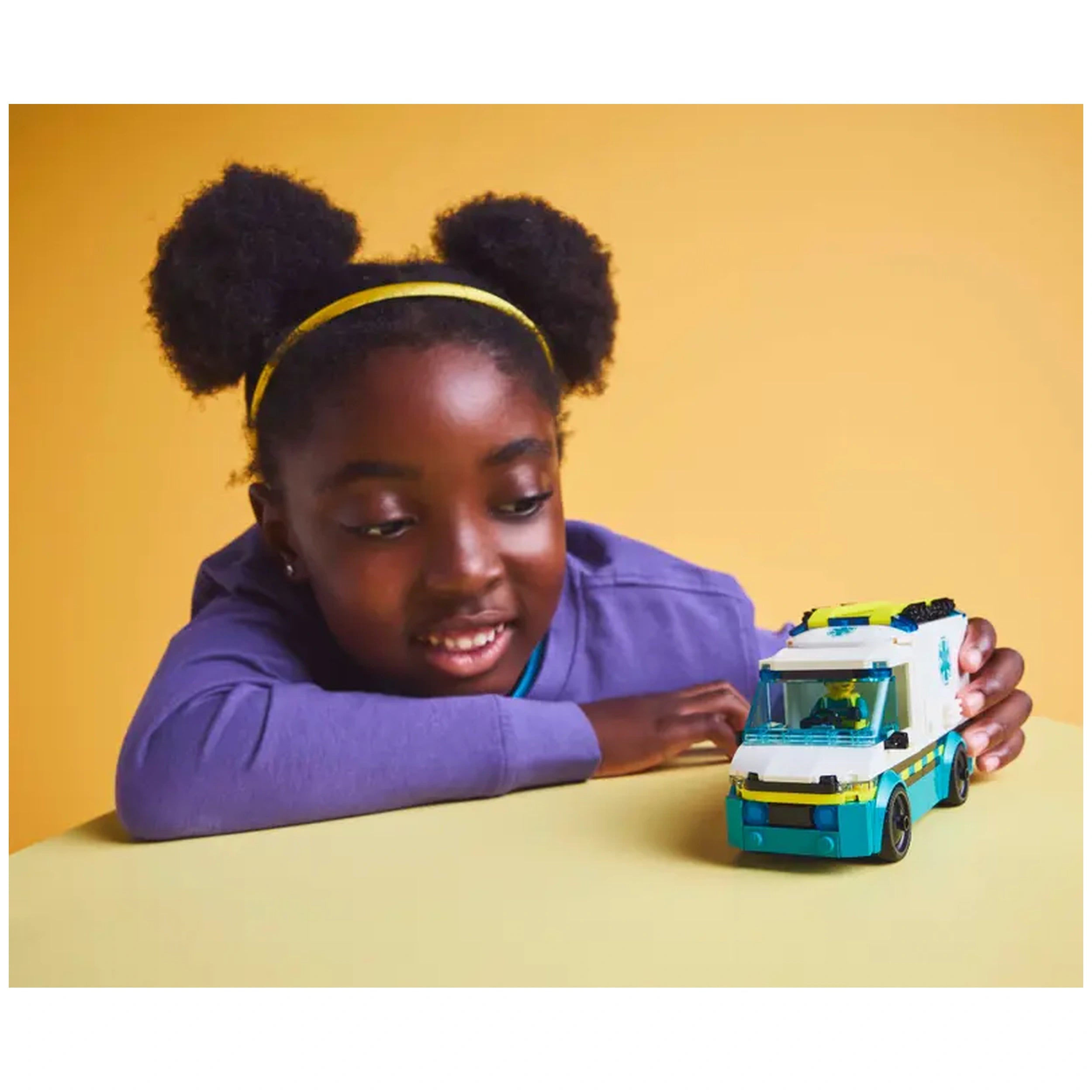 A child with two fluffy pigtails excitedly examines a colorful LEGO® City Emergency Ambulance on a yellow tabletop.