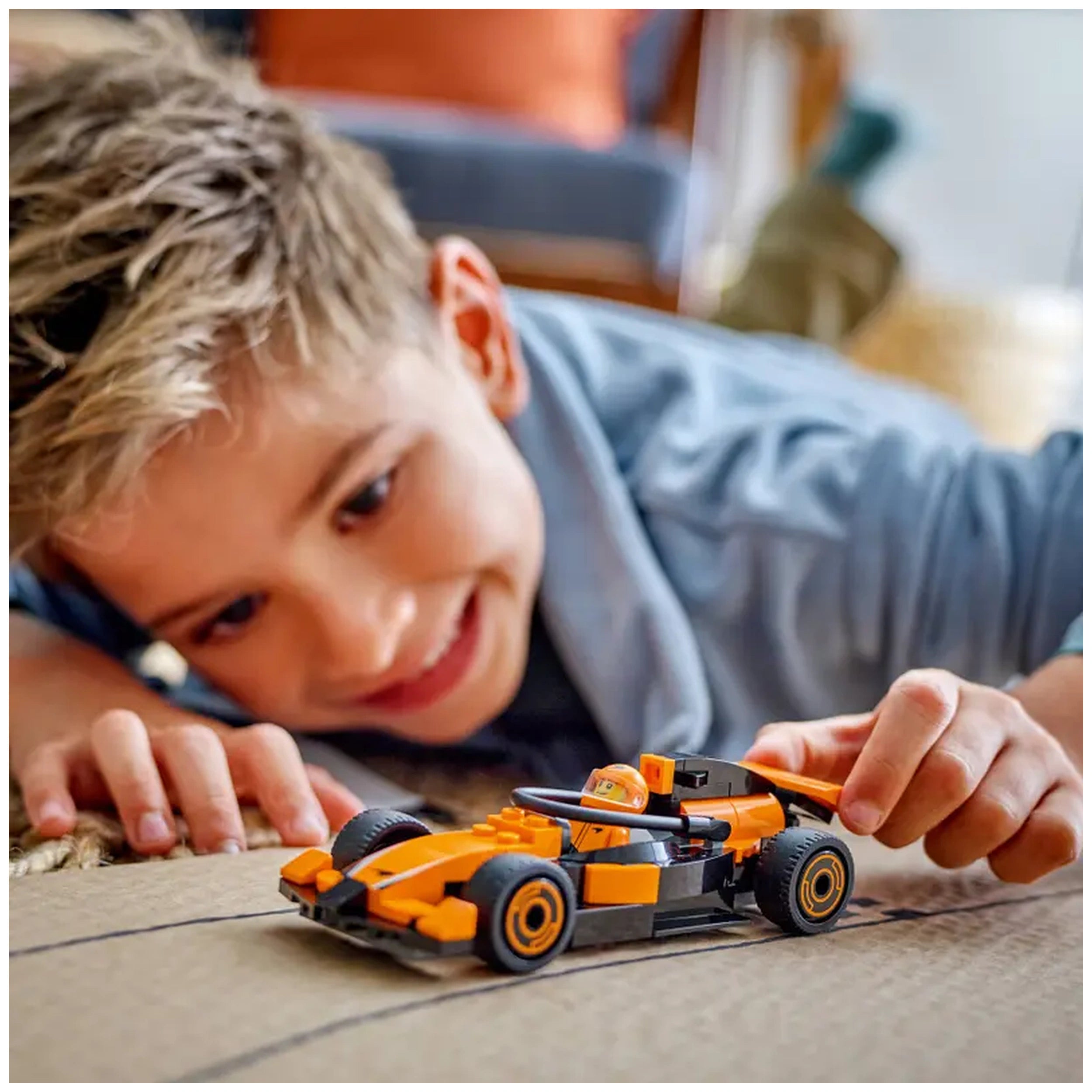 Young boy with short blonde hair excitedly playing with a colorful LEGO® McLaren race car on a cardboard surface.