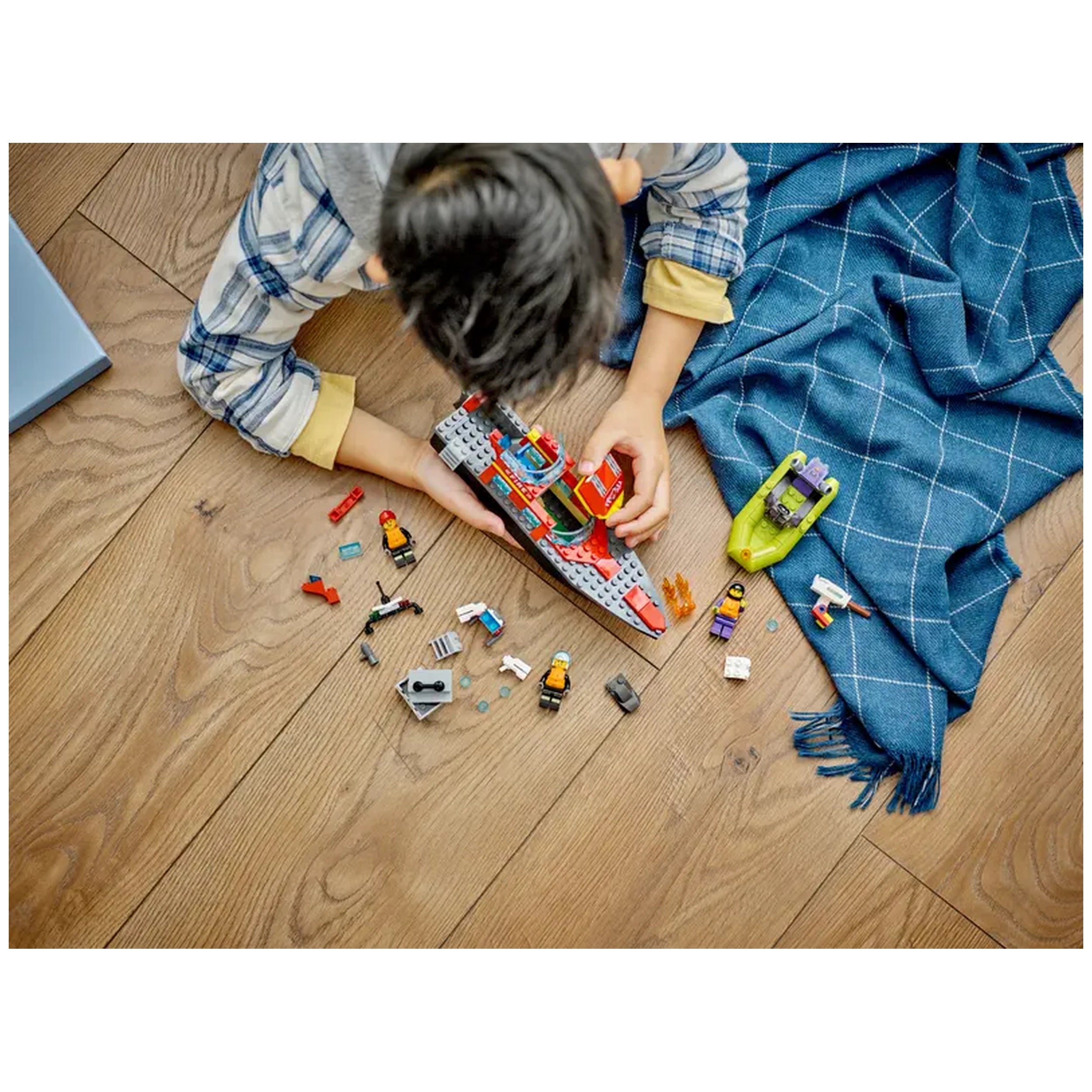 A child builds with the LEGO® City Fire Rescue Boat set, surrounded by various colorful pieces and minifigures on a wooden floor.
