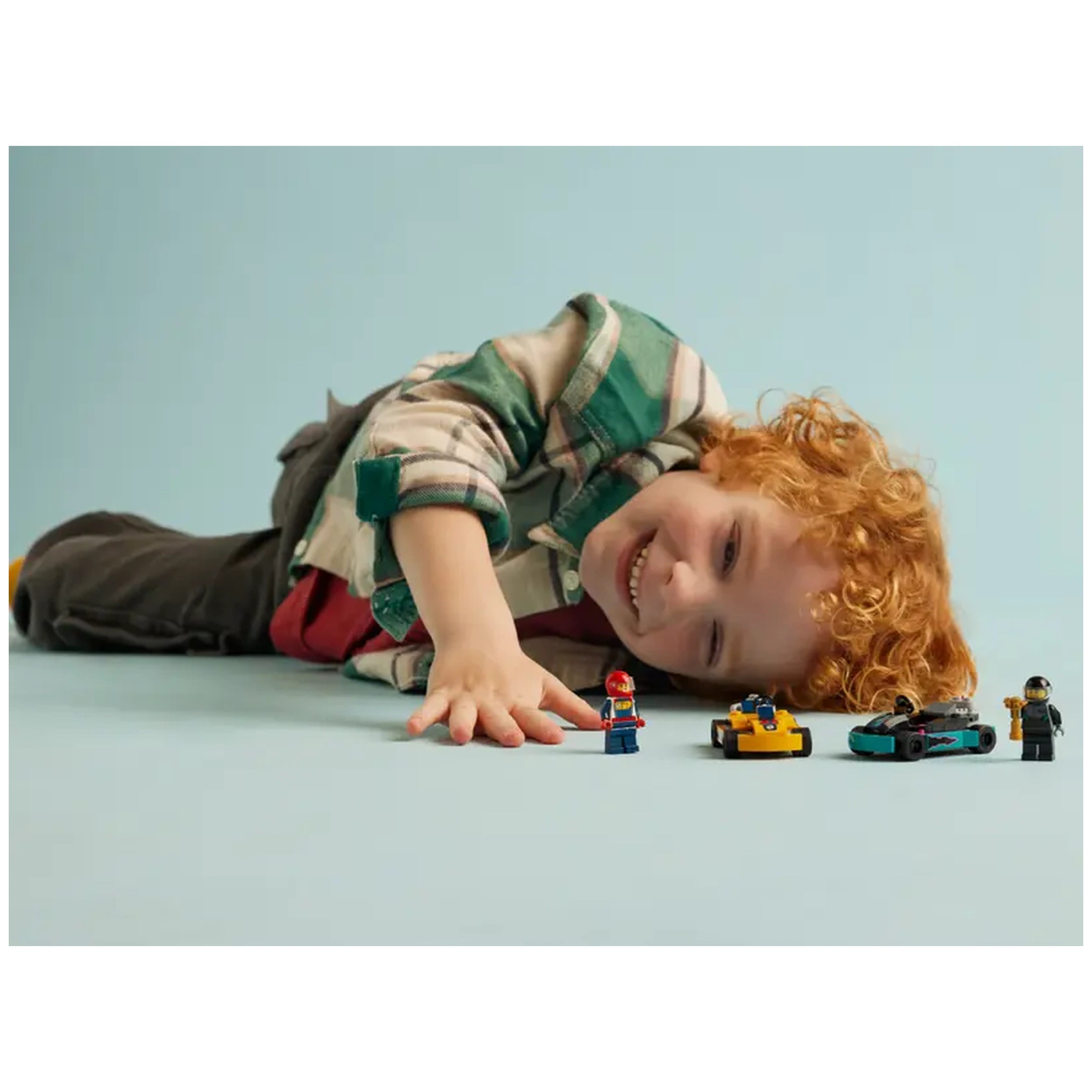 A joyful child with curly red hair lies on a light blue surface, reaching towards two mini LEGO go-karts—one orange and one turquoise—while holding a race driver minifigure. Another minifigure stands beside the go-karts, emphasizing the fun of the LEGO City Go-Karts and Race Drivers set.