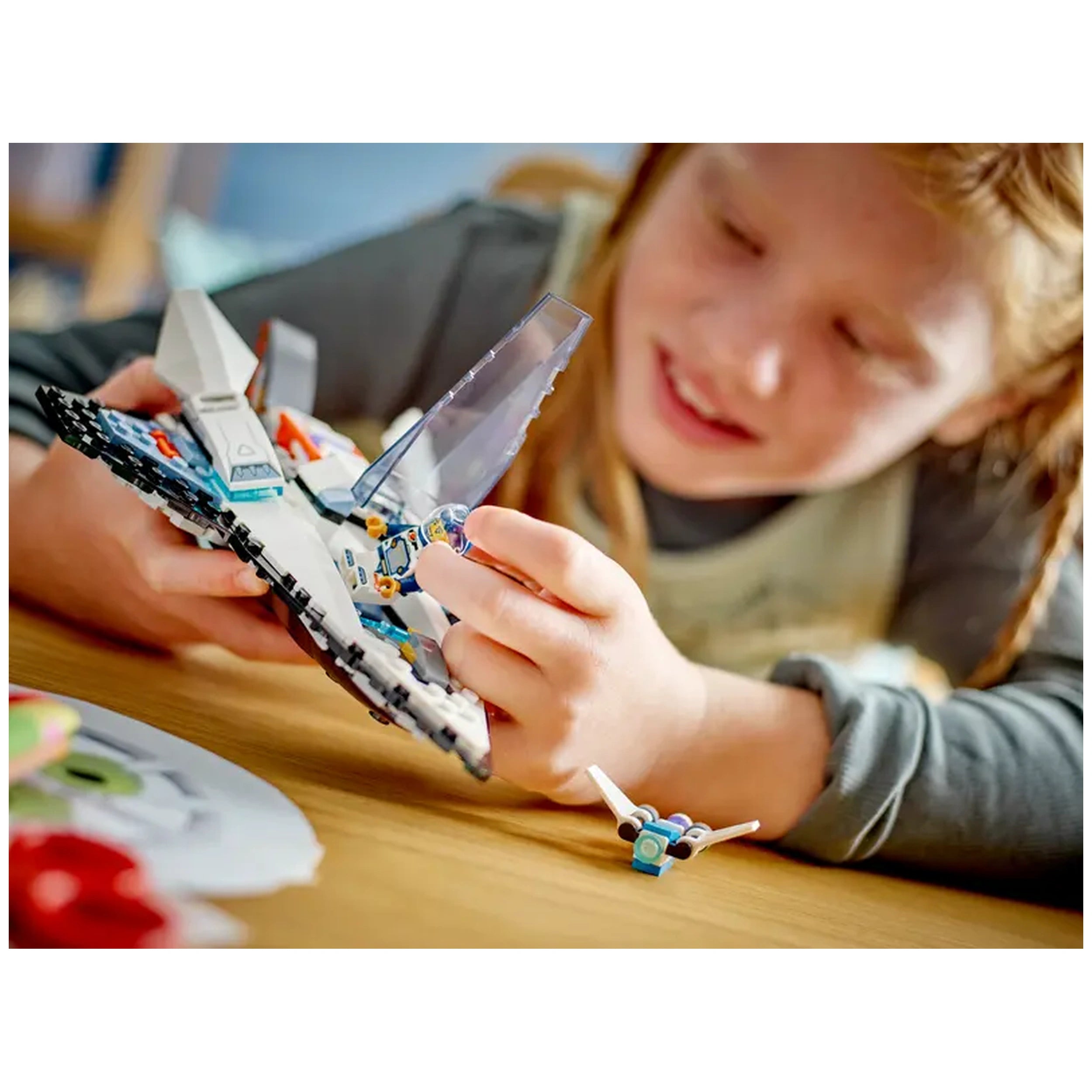A child with long red hair is focused on assembling the LEGO® City Interstellar Spaceship construction set, holding a minifigure. The toy spaceship features a sleek design with blue and white colors, while a small drone bot is on the table nearby, ready for imaginative play.
