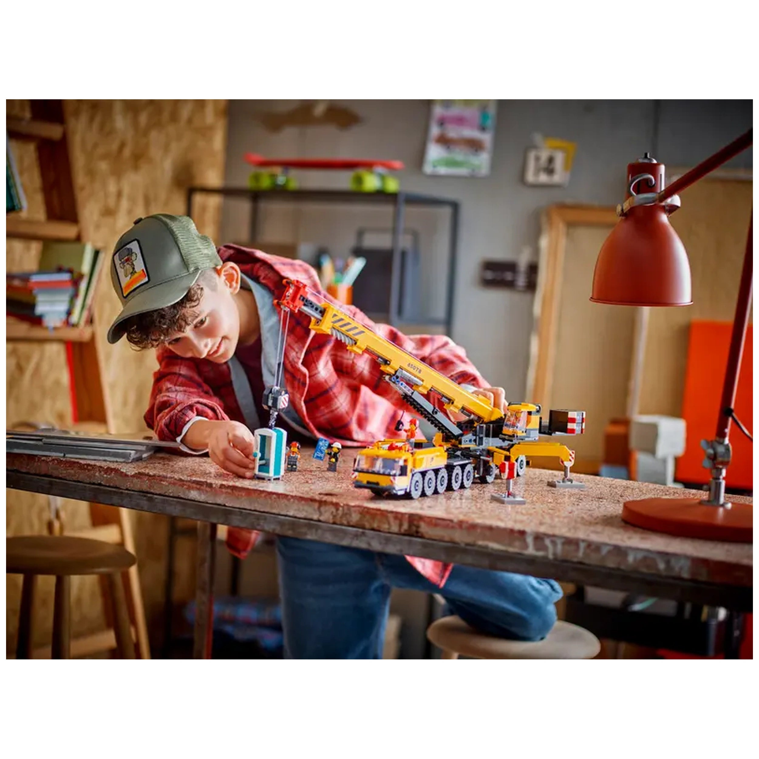 A young boy in a red plaid shirt is playing with a LEGO® City Mobile Construction Crane set on a table, carefully maneuvering a small blue object. The crane's arm is extended, featuring vibrant yellow and orange colors, and the boy's concentration highlights his imaginative play.