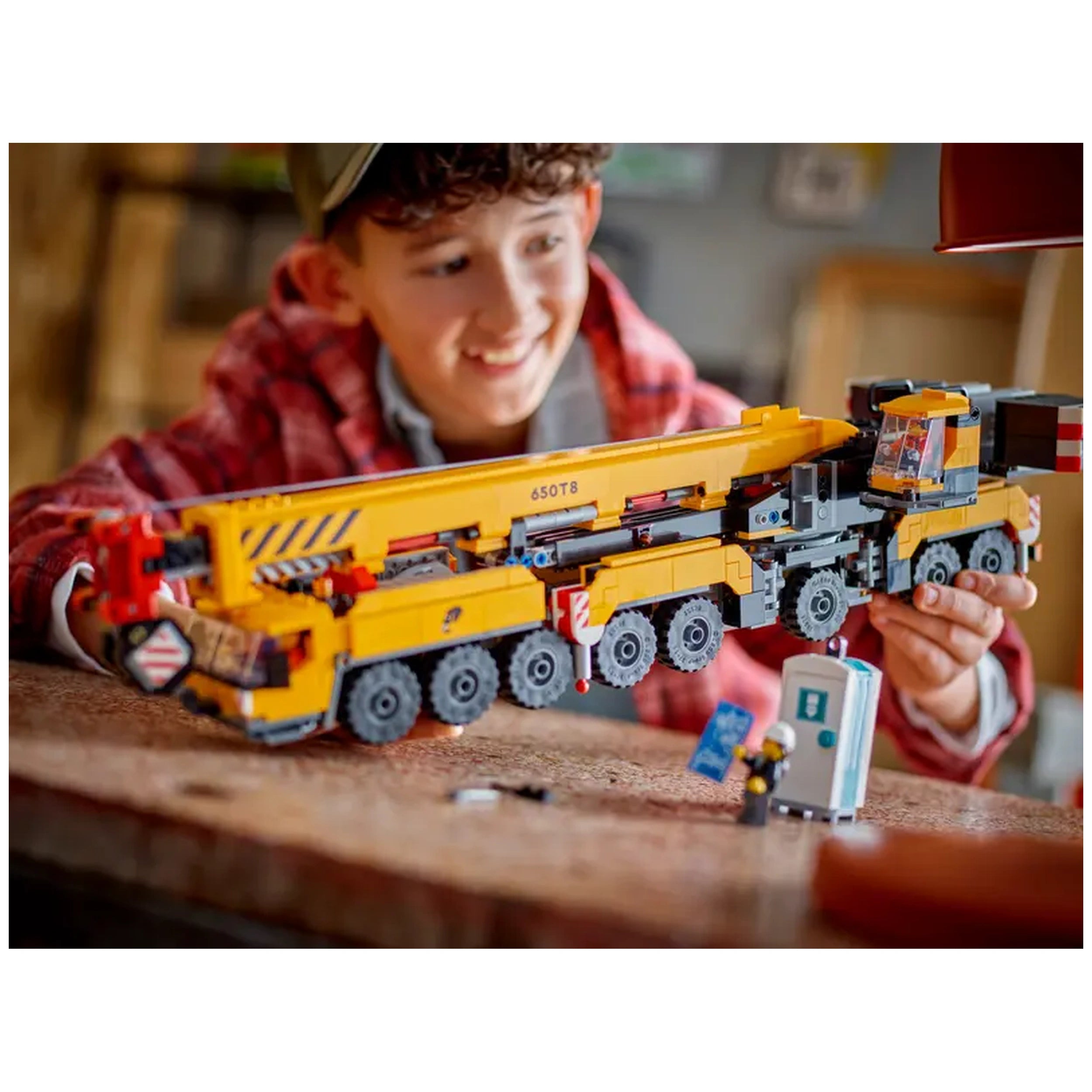 A young boy wearing a plaid shirt smiles while playing with a bright yellow LEGO® Mobile Construction Crane. The detailed model features a long extendable boom and a 16-wheel truck base. Nearby, a miniature construction worker interacts with a blue object, showcasing an imaginative play scene.
