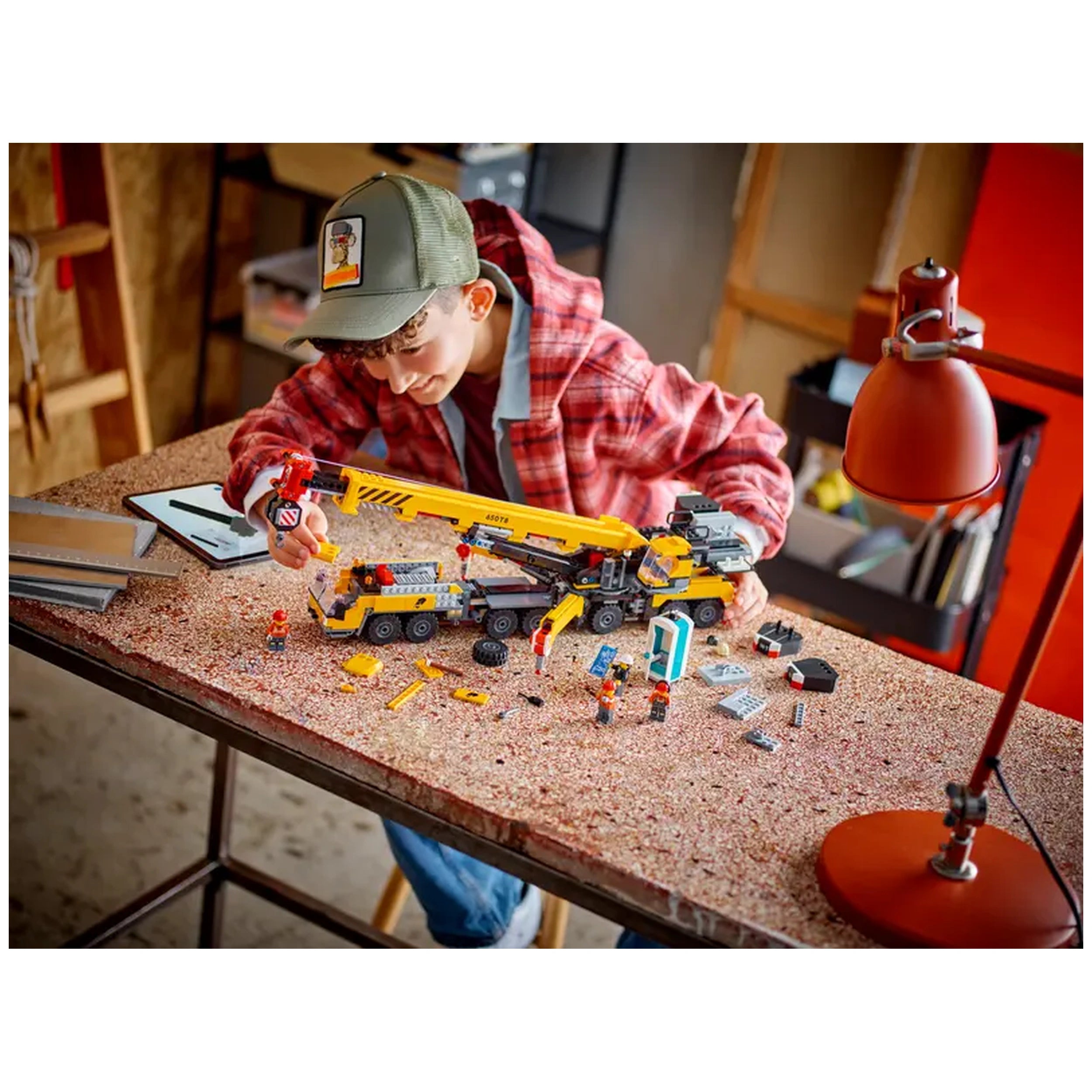 A young boy in a red plaid jacket focuses on assembling the LEGO® City Mobile Construction Crane set on a work table. Bright yellow crane parts and miniature construction workers are scattered around him. A red desk lamp casts warm light over the scene, creating an inviting atmosphere in the background.