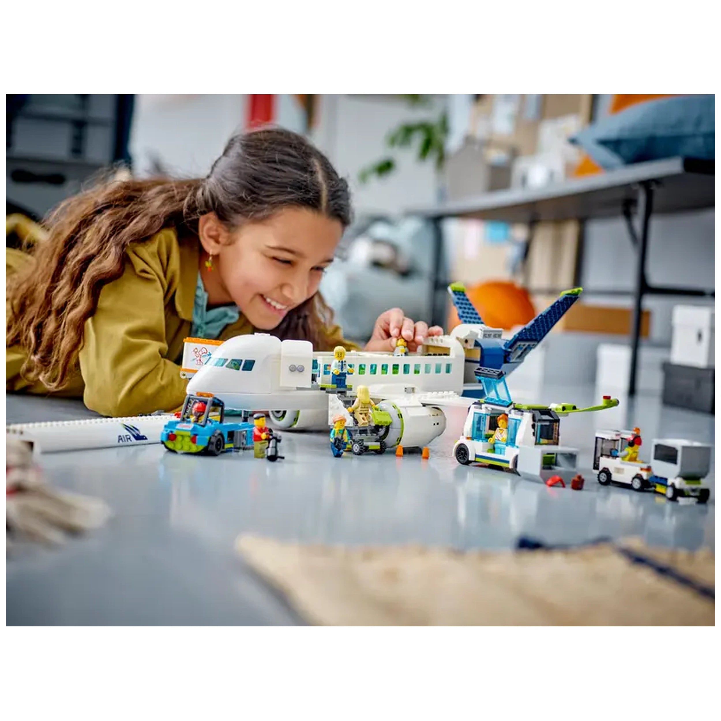 A young girl with curly hair smiles while playing with a LEGO® City Passenger Airplane set, surrounded by detailed minifigures and vehicles.