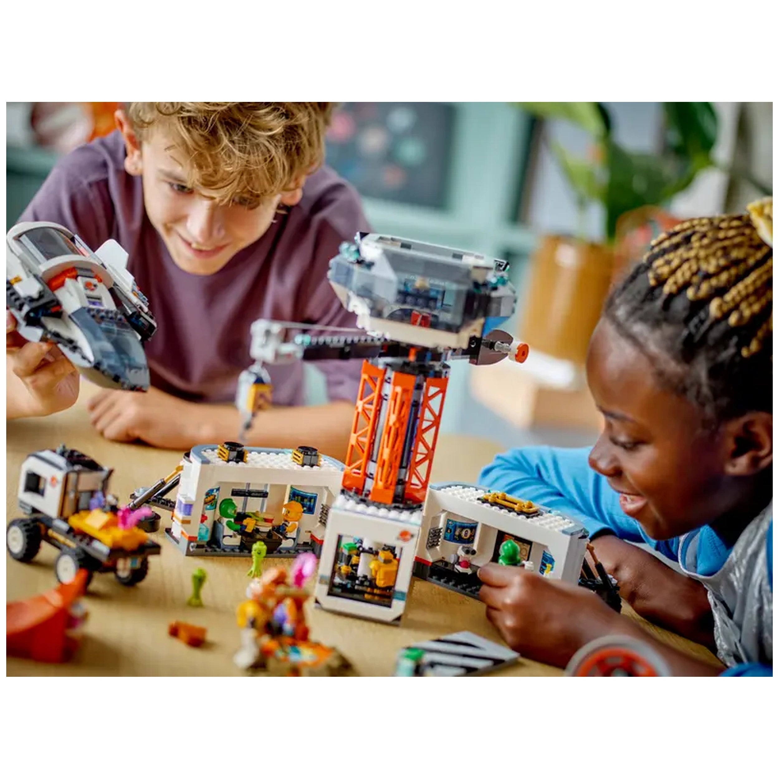 Two children joyfully playing with the LEGO® City Space Base and Rocket Launchpad set, featuring a colorful space crane, construction vehicles, and intricate building elements on a table.