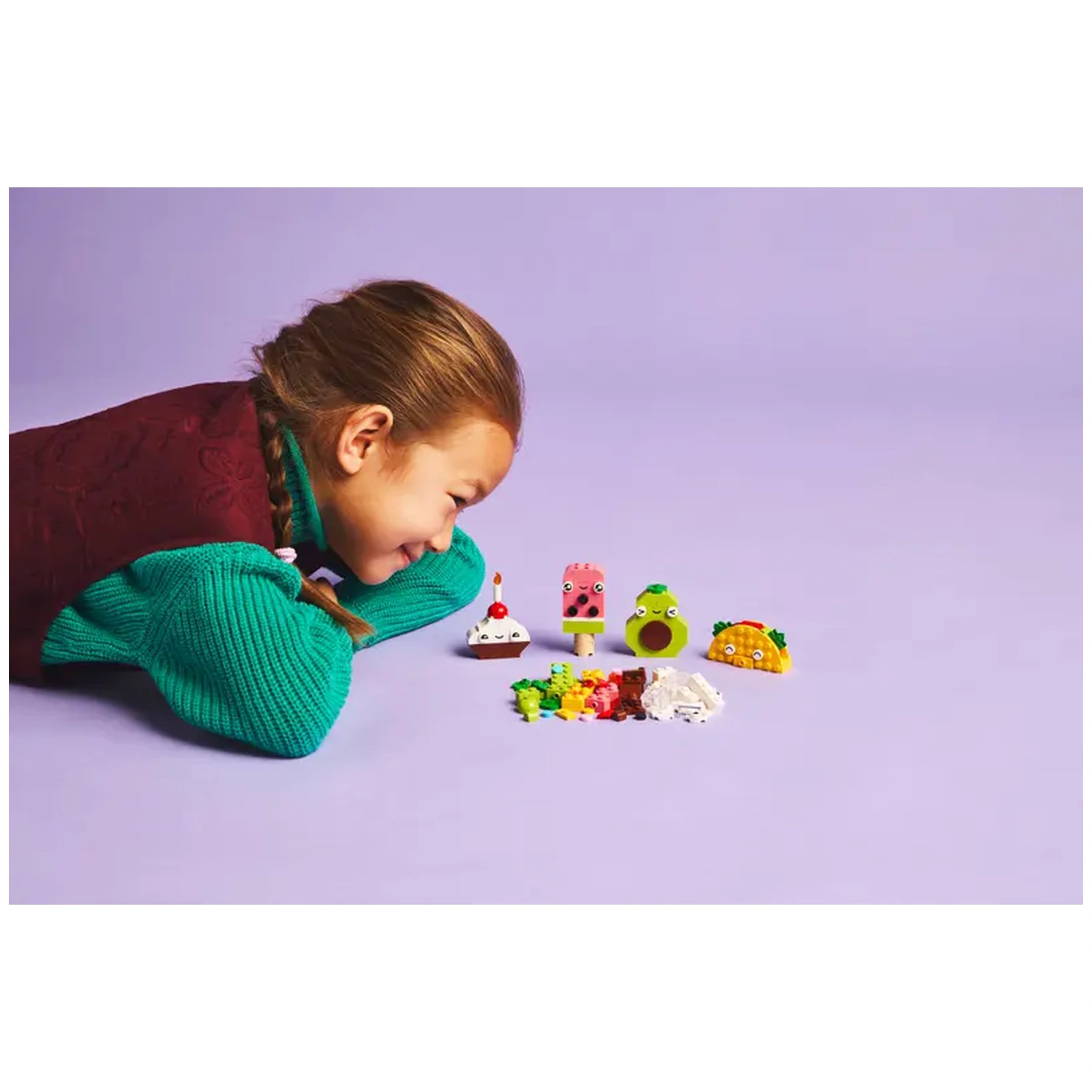 A young girl with braided hair leans over colorful LEGO food-themed characters, including a cupcake, ice cream, avocado, and taco, set against a lilac background.