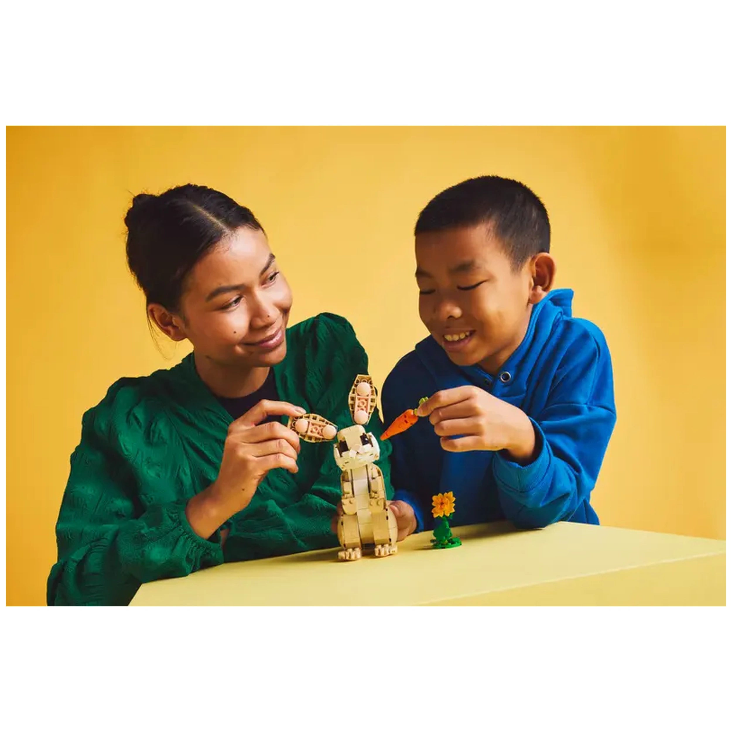 A woman and a boy play with the LEGO Creator Cute Bunny set, featuring a posable bunny, a sunflower, and a carrot against a bright yellow background.