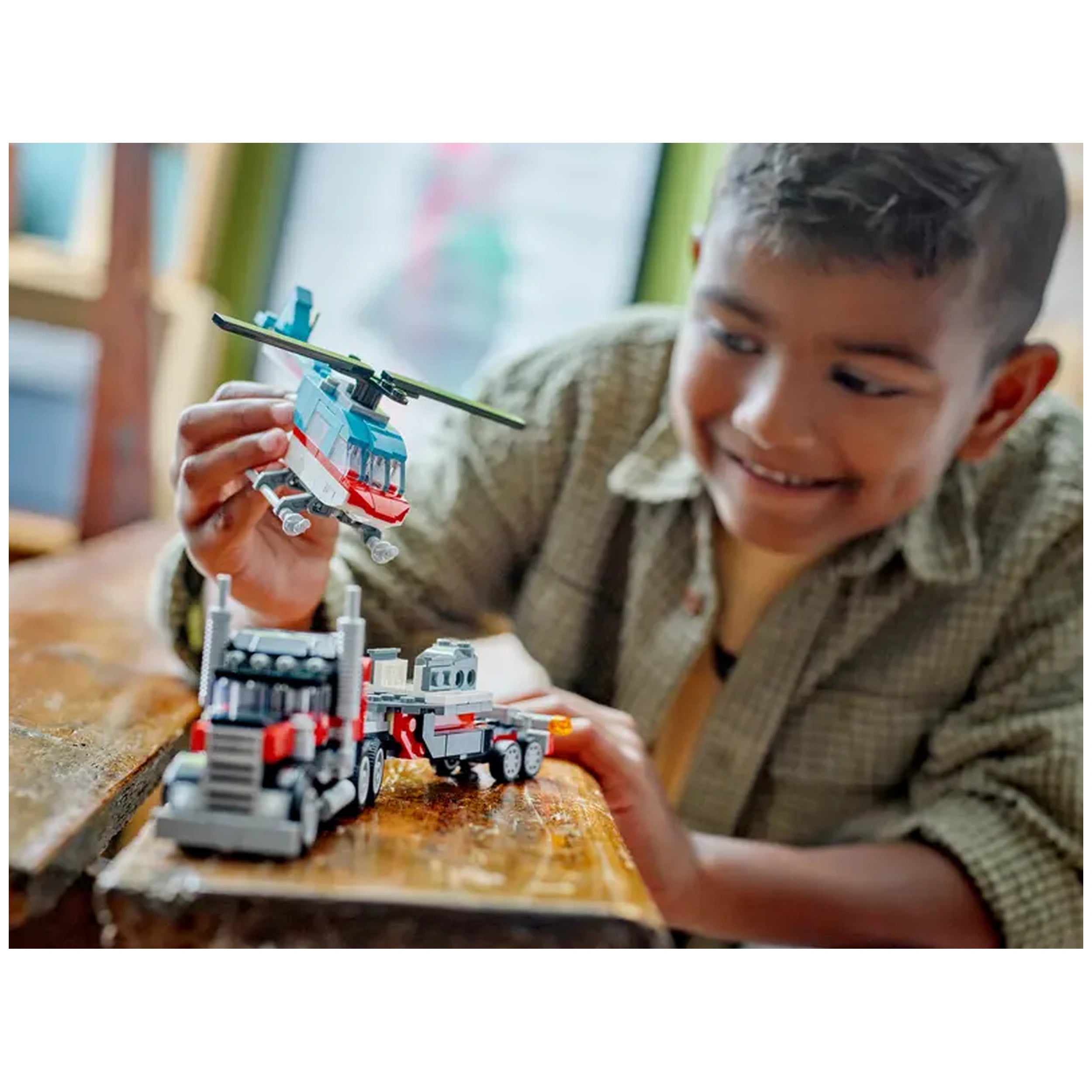 A delighted boy plays with a LEGO® Creator construction set, holding a blue helicopter while examining a flatbed truck on a wooden table.