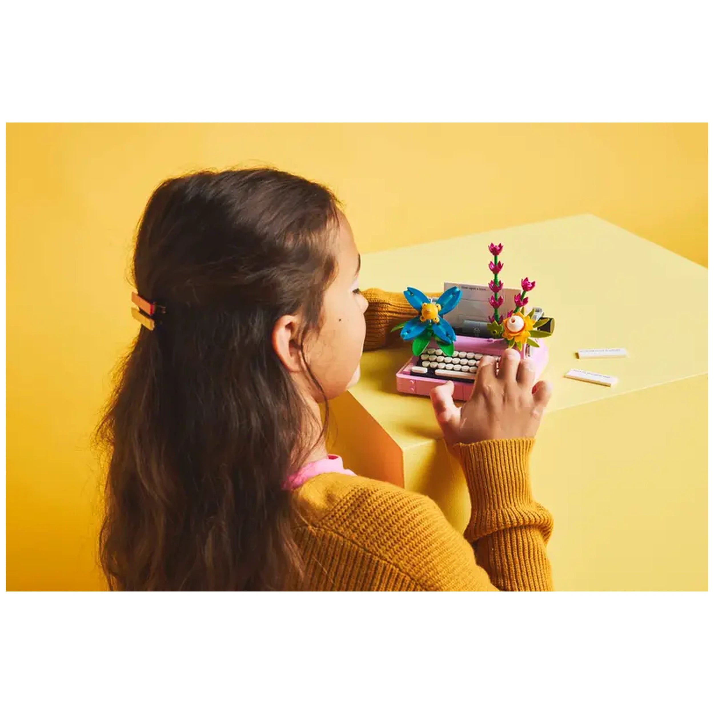 A child with long hair, wearing a cozy brown sweater, engages with a LEGO® Creator typewriter set on a bright yellow table. The typewriter features colorful brick-built flowers and a moving keyboard.
