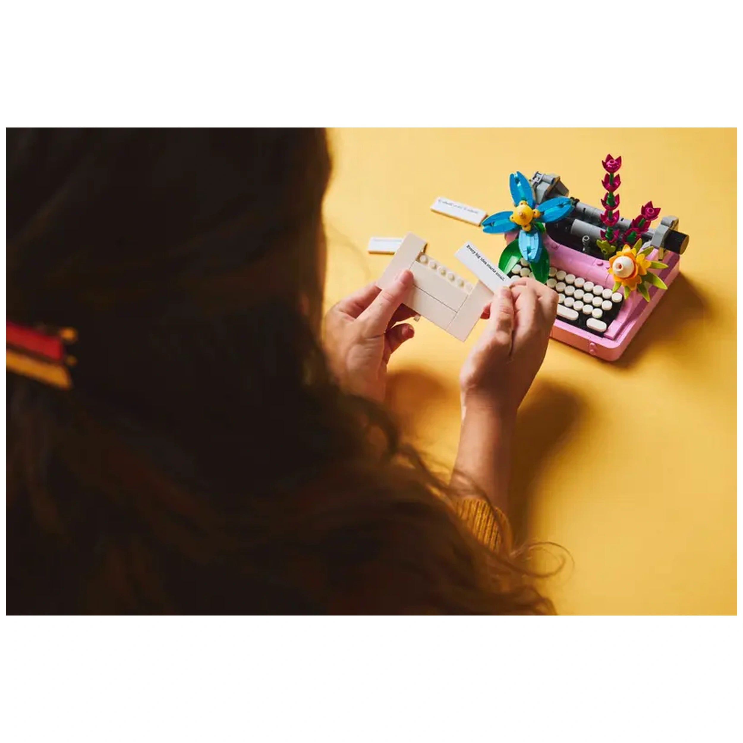 A child holds a brick-built sheet of paper in front of a colorful LEGO® Typewriter, featuring vibrant flowers on a bright yellow background.